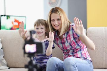 Joyful mother and daughter hold their hand in greeting to video camera screen. Family video blogging concept