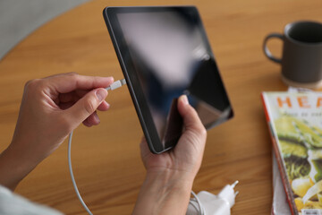 Woman connecting charger cable to tablet at wooden table, closeup
