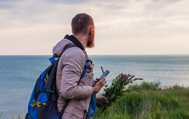 Bearded man in raincoat with backpack on sea landscape with blue cloudy sky background with smartphone and wild flowers. Guy photographing nature on cell phone, active lifestyle travel concept.