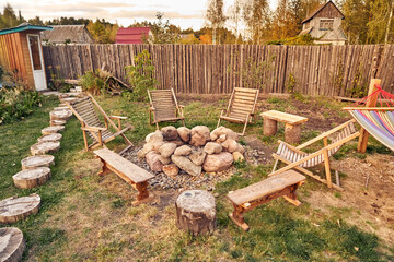 Arranged campfire place in the backyard of the cottage. Wooden sun loungers and benches by the large stone fireplace.