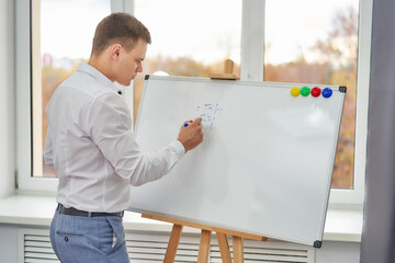 A young man in a white shirt in the office by the window writes with a marker on a flipchart.