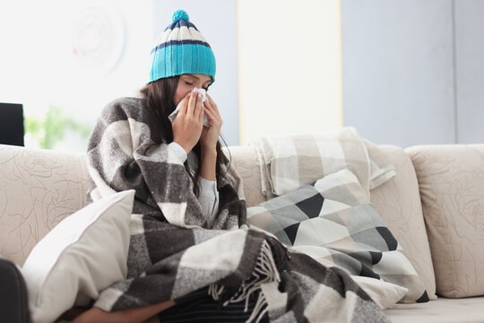 chilled woman on sofa in blanket and hat holds handkerchief. Seasonal colds concept