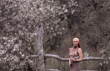 the girl is standing near a wooden fence and a blooming spring tree, dressed in a brown scarf tied on her head, a brown polka-dot T-shirt, a skirt, holding a sprig of cherry blossoms