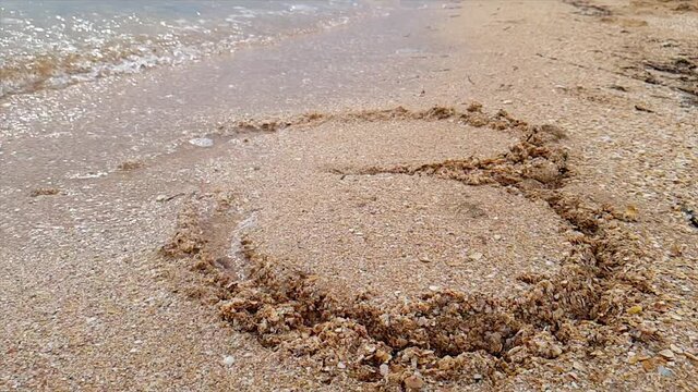 The wave washes away the heart nestled in the sand. Woman draws a heart in the sand. A woman draws a heart on the seashore which is washed away by a wave.