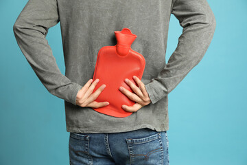 Man using hot water bottle to relieve back pain on light blue background, closeup