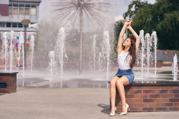 Beautiful young woman with brown hair sitting on a bench near the city fountain, girl raises her hands up, happiness concept.