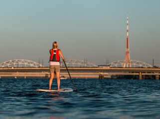 Young woman rowing with SUP stand up paddle boards along the river, in the background TV tower and bridges over Daugava river in Latvia