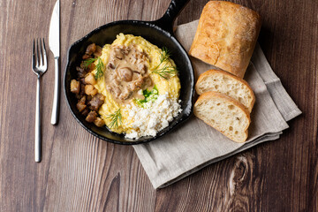 Banosh - a ukrainian dish made of cornmeal. Served in black pan with cracklings, mushrooms and hutsul cheese. Close-up isolated on a wooden background with cutlery, sliced bread and grey linen napkin.