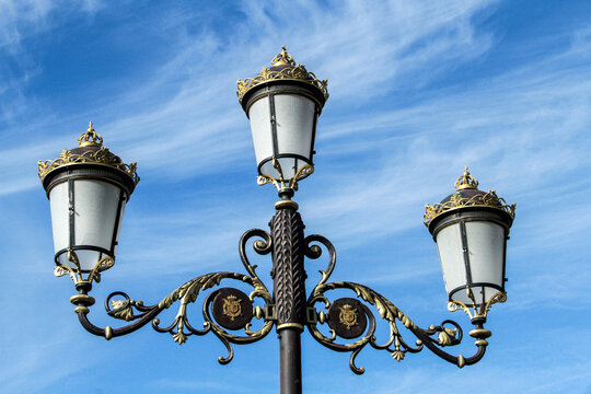 Old vintage black decorative lantern with the royal coat of arms and white frosted glass on the pole. Three street lamps on one pole. Sunny blue sky with white clouds