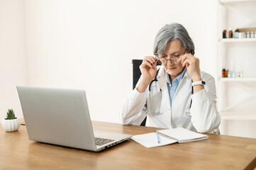 Senior old female doctor with stethoscope in a lab coat and glasses is done with work, removing her...