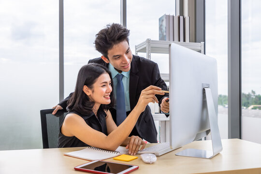 Asian Businessman And Businesswoman Discussing With Computer At Desk, Pointing At Monitor Screen, Working Together At Workplace Office.