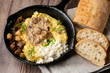 Banosh - a ukrainian dish made of cornmeal. Served in black pan with cracklings, mushrooms and hutsul cheese bryndza. Close-up isolated on a wooden background with sliced bread and grey linen napkin.
