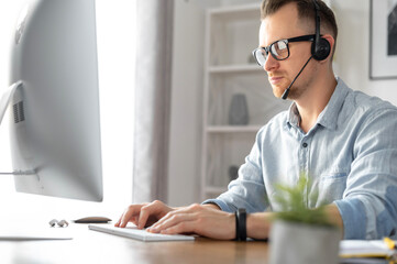 Side view of young male wearing formal shirt using a headset and contemporary PC in the office, intelligent guy talking online and typing sitting at the desk. Call center, help line, support service