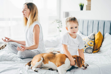 Mother doing morning exercises in yoga pose while her little daughter playing near at home.