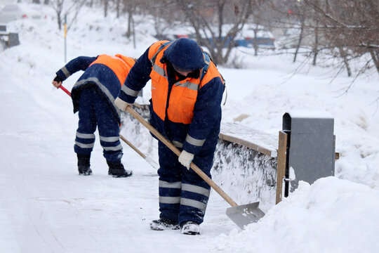 Snow removal in winter city, street cleaning at snowfall. Two communal services workers in orange uniform with a shovel clears snow on a sidewalk