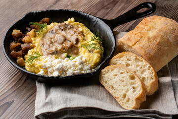 Banosh - a ukrainian dish made of cornmeal. Served in black pan with cracklings, mushrooms and hutsul cheese bryndza. Close-up isolated on a wooden background with sliced bread and grey linen napkin.