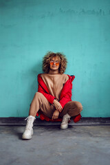 Young fashionable woman with curly hair, in red jacket crouching in front of the wall and looking...