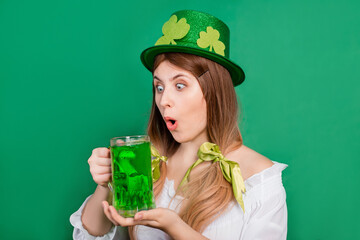 Woman in festive saint patrick's day costume and hat decorated with shamrock holding a mug of green beer, isolated on green background.