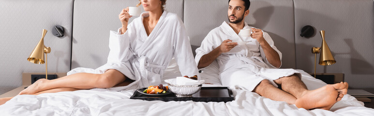 Muslim man holding coffee cup near girlfriend and breakfast on hotel bed, banner