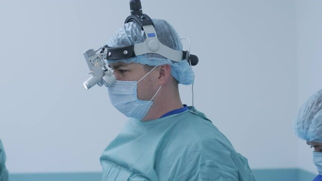 The surgeon prepares for the operation, fastens the sterile gown in the modern operating room. Doctor is equipped in mask, medical headlamp, surgical loupes
