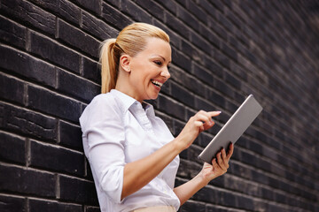 Attractive innovative smiling blond fashionable businesswoman standing in front of black wall and using tablet.