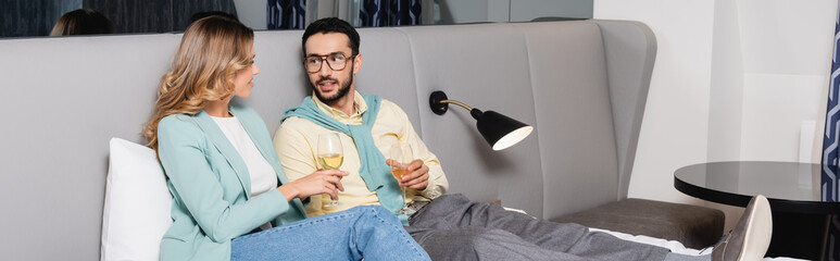 Interracial couple holding glasses of wine on bed in hotel room, banner