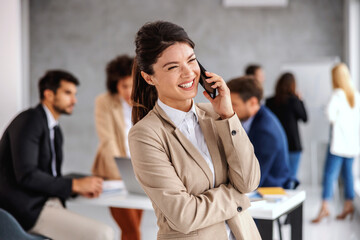 Smiling cheerful young businesswoman standing in boardroom and talking on the phone. In background are her colleagues working on important project.
