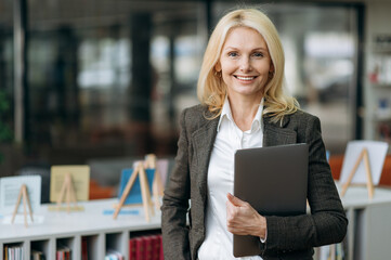 Portrait of influential elegant businesswoman with blonde hair in stylish formal suit. Happy successful middle aged lady looks directly at the camera, friendly smiling, holding laptop in arms