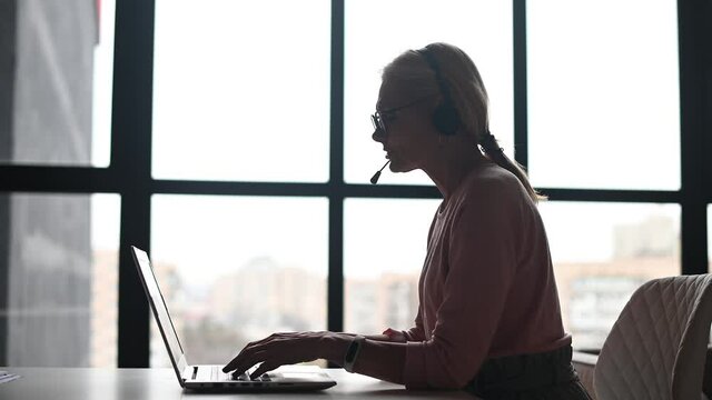 A silhouette of an elegant mature blonde woman or middle-aged customer service representative sitting at the desk against panoramic window, wearing headset, glasses, looking at the laptop and talking