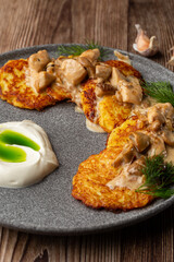 Fried potato pancakes served with mushrooms, sour cream and dill on a grey round plate. Close-up isolated dish on wooden background with cloves of garlic on the table.
