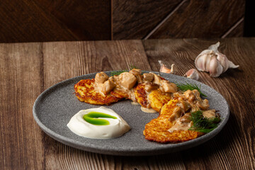 Fried potato pancakes served with mushrooms, sour cream and dill on a grey round plate. Close-up isolated dish on wooden background with cloves of garlic on the table.