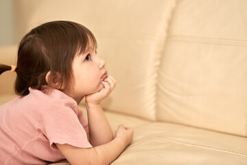 A little girl is lying on her stomach on the couch. She rested her chin on her hand. She has a dreamy, thoughtful look. Selective focus. Copy space.