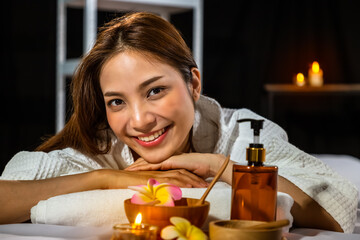 Obraz na płótnie Canvas Beautiful young Asian woman resting on bed in spa, with massage oil, scrub powder, and candle, looking at camera.
