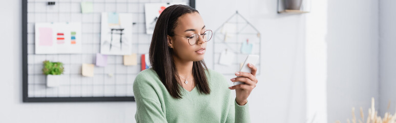 young african american architect in eyeglasses messaging on smartphone, banner