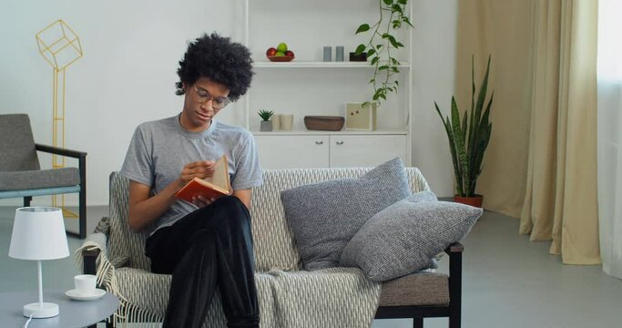 Afro american guy teen student wears glasses and casual clothes sits on sofa in modern living room holds orange hardback book in his hands reads page studies literature enjoys textbook during lockdown