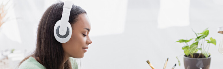 Naklejka premium side view of smiling african american interior designer listening music in wireless headphones, banner