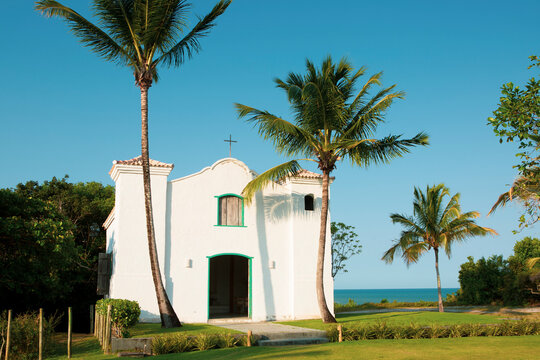 Rustic small church or chapel exterior with coconut trees in front of it and a metal cross on the roof under blue sky in a sunny day in Bahia, Brazil.