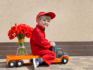 funny cute 4 years old boy in a red mechanic overalls and a cap sitting on a toy truck with large...