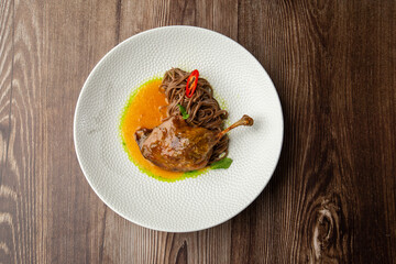 Roasted duck thigh with brown spaghetti. Served with yellow sauce and decorated with mint leaves and slice of chilli pepper. Close-up in a white bowl on wooden background. Top view, copy space.