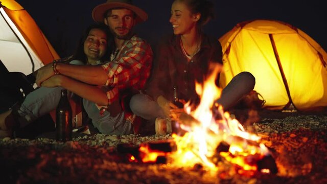 group of friends relaxing at night on beach camping at tents, sitting at bonfire with beer, talking and enjoying warm of fire. summer traveling together with tents