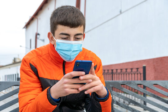 Stock photo of young boy wearing face mask due to covid19 typing on his phone while sitting in a bench in the street.