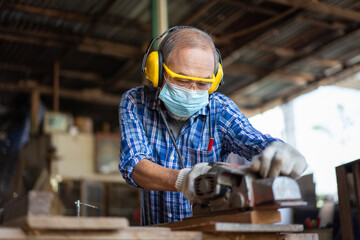 Asian senior carpenter wear hygienic mask protect using electric planer adjust surface on wooden plank at the carpentry workshop
