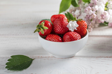 close-up of red strawberries in a white bowl on a light wooden background with flowers