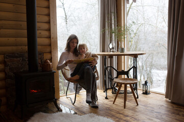 Mother and cute toddler child in a little fancy wooden cottage, reading a book, drinking tea