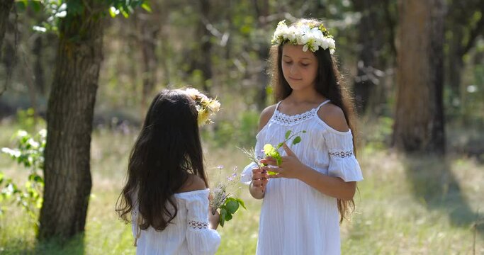 Close-up portrait of  Caucasian teenager girl with long brown hair and blue eyes in flower wreath looking at her younger sister gathering bouquet on nature background. 4k 50 fps slow motion