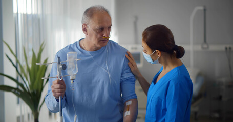 Aged man patient with dropper talking to nurse standing in hospital ward