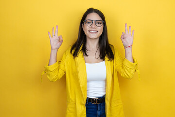 Young brunette businesswoman wearing yellow blazer over yellow background doing ok sign with fingers and smiling, excellent symbol