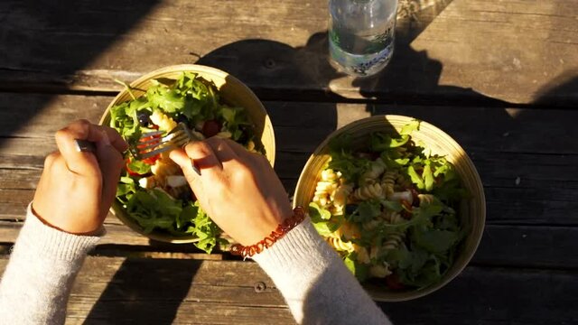Caucasian woman mixing a mediterranean greek salad with feta cheese, olives, cherry tomatoes, rucola and fusilli pasta on a bamboo bowl in slow motion