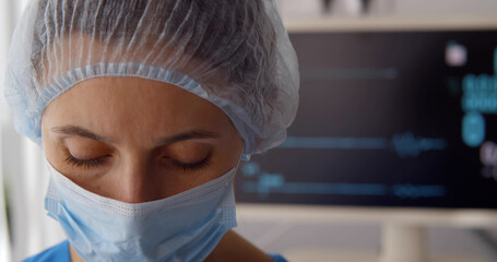 Fototapeta na wymiar Sad doctor in safety mask and hat standing near monitor with no heart rate