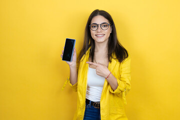 Young brunette businesswoman wearing yellow blazer over yellow background holding smartphone showing screen smiling happy pointing with hand and finger